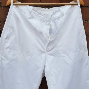 May include: White cotton pants with a button fly and a single front pocket. The pants are hanging on a wooden hanger.