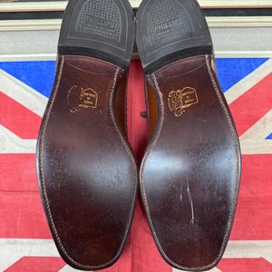 May include: A pair of brown leather dress shoes, soles facing up, with the text "PHILLIPS CUSHION NO MARK" and "MADE IN ENGLAND" visible. The shoes are set against a Union Jack flag.