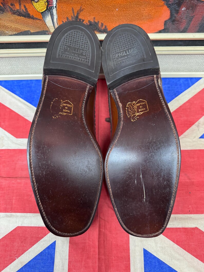 May include: A pair of brown leather dress shoes, soles facing up, with the text "PHILLIPS CUSHION NO MARK" and "MADE IN ENGLAND" visible. The shoes are set against a Union Jack flag.