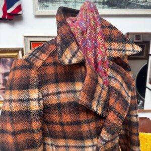 May include: A vintage plaid coat in shades of brown and orange with a wide collar. A paisley scarf in red, purple, and yellow is draped over the collar. The coat has a classic, tailored design, suggesting a retro style.