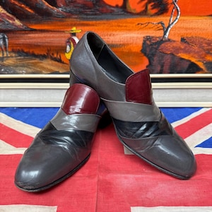 K&ouml;nnte beinhalten: Ein Paar Vintage-Lederschuhe in Grau-, Schwarz- und Burgundert&ouml;nen. Die Schuhe haben ein einzigartiges geometrisches Design mit einem niedrigen Absatz. Die Schuhe werden auf einer Union Jack-Flagge mit einem Landschaftsbild im Hintergrund pr&auml;sentiert.