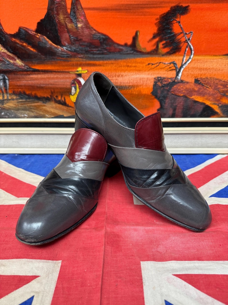 K&ouml;nnte beinhalten: Ein Paar Vintage-Lederschuhe in Grau-, Schwarz- und Burgundert&ouml;nen. Die Schuhe haben ein einzigartiges geometrisches Design mit einem niedrigen Absatz. Die Schuhe werden auf einer Union Jack-Flagge mit einem Landschaftsbild im Hintergrund pr&auml;sentiert.