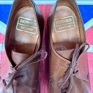 May include: A pair of brown leather dress shoes with laces, featuring the brand name "Hornes London" printed in gold on the inner sole. The shoes are set against a backdrop of the Union Jack flag.