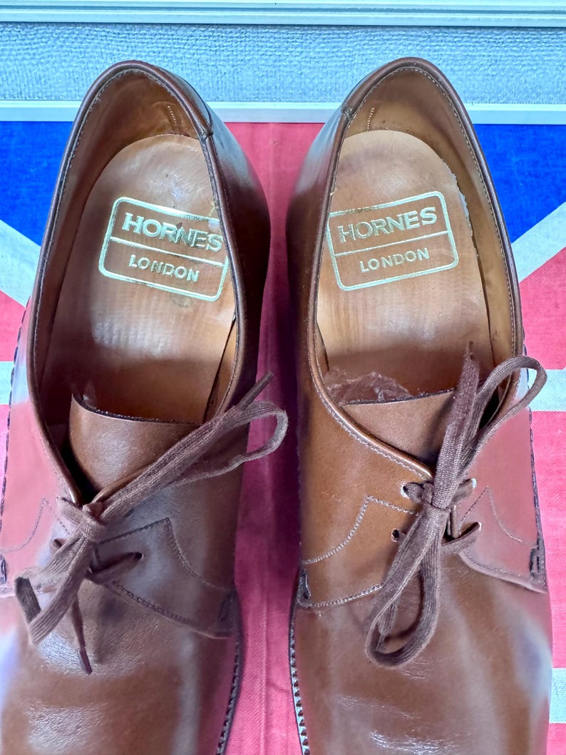 May include: A pair of brown leather dress shoes with laces, featuring the brand name "Hornes London" printed in gold on the inner sole. The shoes are set against a backdrop of the Union Jack flag.
