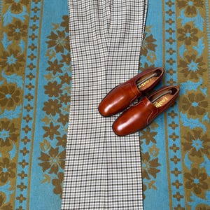 May include: A pair of brown leather loafers and a pair of checkered trousers are displayed on a patterned rug. The trousers have a black, white, and tan checkered pattern. The loafers are a rich brown colour.