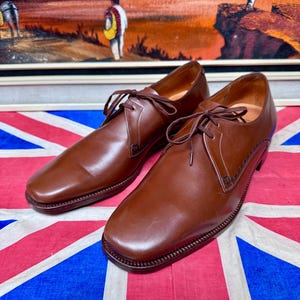 May include: A pair of brown leather dress shoes with laces, featuring a square toe design. The shoes are displayed on a Union Jack flag, with a painting in the background.