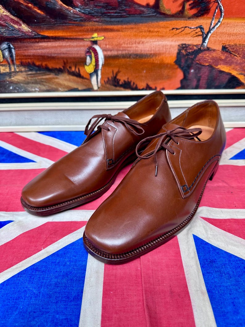 May include: A pair of brown leather dress shoes with laces, featuring a square toe design. The shoes are displayed on a Union Jack flag, with a painting in the background.