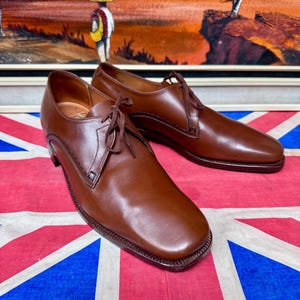 May include: A pair of brown leather dress shoes with a square toe and lace-up closure. The shoes have a stitched design along the edges and are displayed on a Union Jack flag.