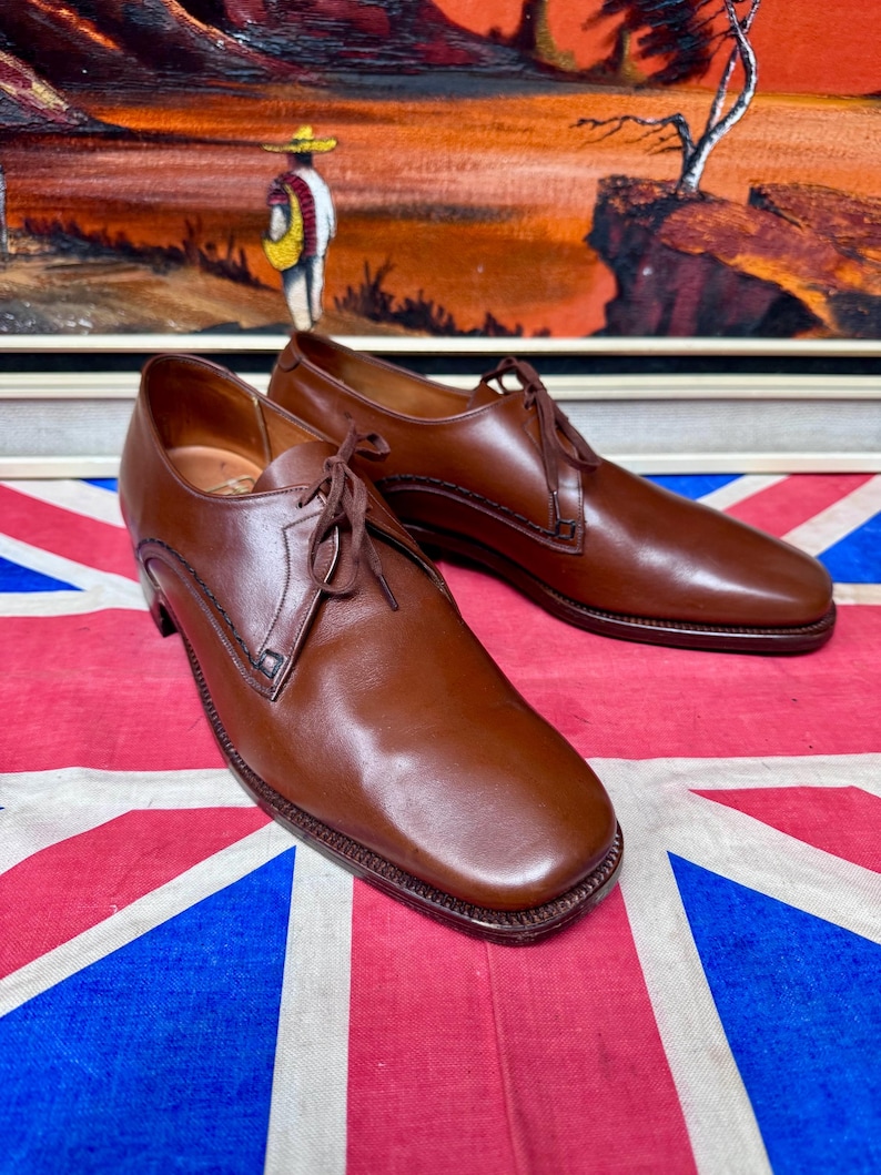 May include: A pair of brown leather dress shoes with a square toe and lace-up closure. The shoes have a stitched design along the edges and are displayed on a Union Jack flag.