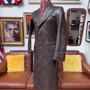 Pode incluir: Um casaco de couro castanho vintage com um design clássico, com gola entalhada, frente abotoada e bolsos. O casaco é exibido em um manequim de madeira. Um lenço colorido é drapeado ao redor do pescoço do manequim.