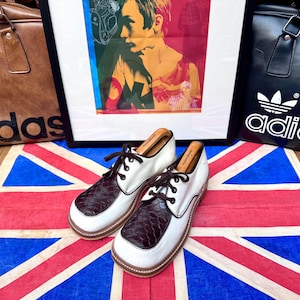 May include: A pair of white and brown leather shoes with dark brown laces, displayed on a Union Jack flag. The shoes have a textured brown toe cap. Two Adidas bags and a framed print are in the background.