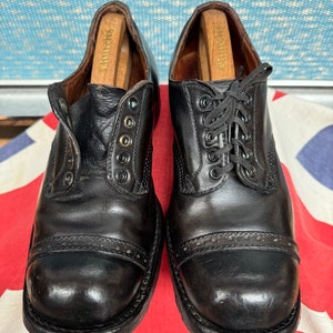 May include: A pair of black leather shoes with wooden shoe trees. The shoes feature a cap toe design and are laced with black laces. The shoes are displayed on a Union Jack flag.
