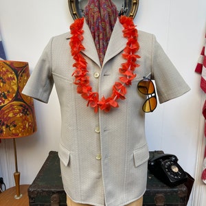 May include: A light gray short-sleeved blazer with four buttons and a patch pocket on the left side. The blazer is worn on a mannequin with a red flower lei and a paisley scarf.