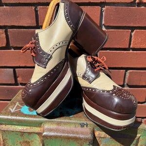 May include: A pair of two-tone platform oxford shoes featuring cream and dark brown leather panels with decorative brogue perforations. The shoes have thick stacked platform soles, chunky block heels, and thin orange laces tied at the front.
