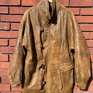 May include: A brown leather jacket with a high collar, a zipper closure, and toggle closures. The jacket has a large pocket on the front and a smaller pocket on the left sleeve.