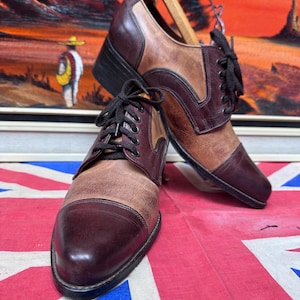 May include: A pair of two-tone leather dress shoes with black laces. The shoes feature a combination of brown and burgundy leather. The shoes are displayed on a Union Jack flag.