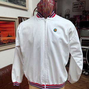Peut inclure: Veste blanche zippée avec un emblème rond sur la poitrine. La veste présente des bordures rayées rouges, blanches et bleues sur le col, les poignets et l'ourlet. La veste est présentée sur un mannequin.