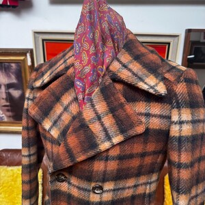 May include: A vintage double-breasted coat in a plaid pattern of orange, brown, and black. The coat has a wide collar and three visible buttons. A paisley scarf in shades of red, purple, and yellow is draped over the coat's collar.