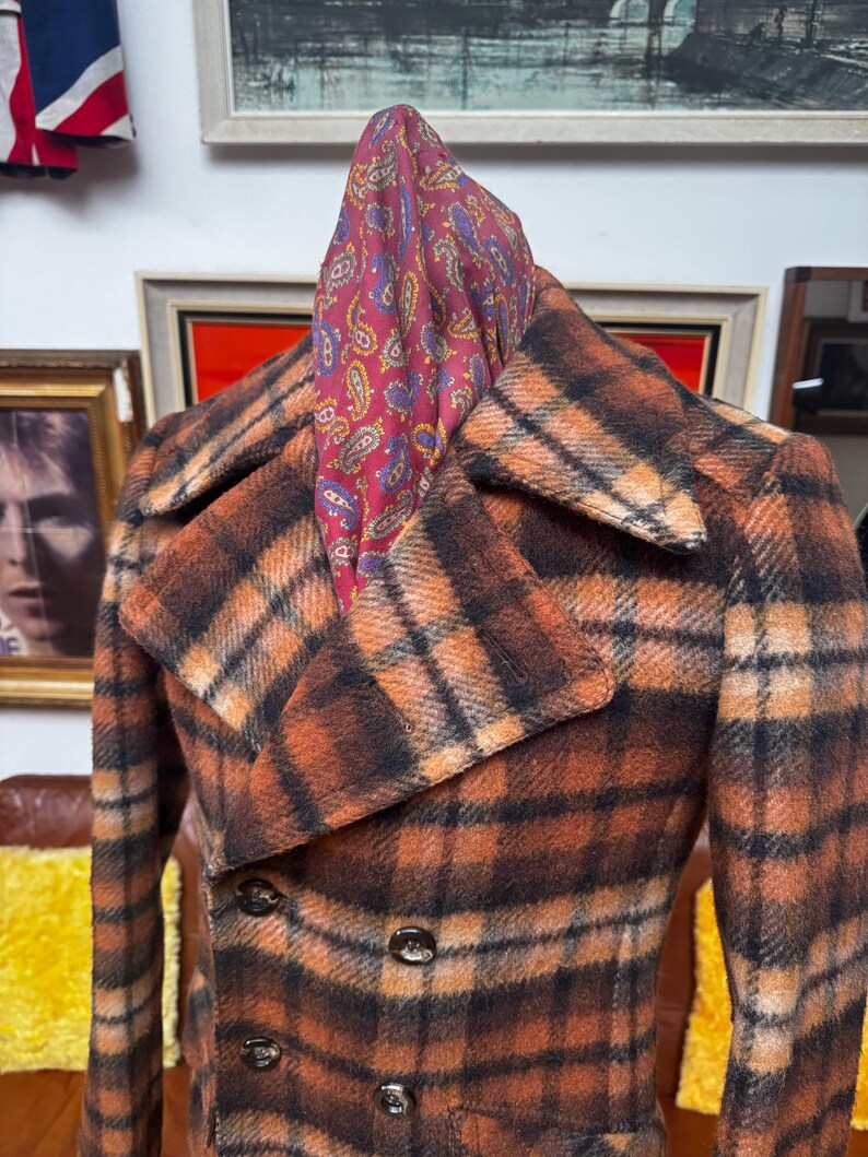 May include: A vintage double-breasted coat in a plaid pattern of orange, brown, and black. The coat has a wide collar and three visible buttons. A paisley scarf in shades of red, purple, and yellow is draped over the coat's collar.