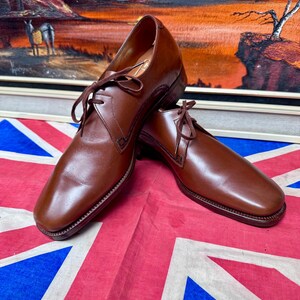 May include: A pair of brown leather dress shoes with laces. The shoes have a square toe and are displayed on a Union Jack flag. The background features a framed landscape painting with a sunset scene.
