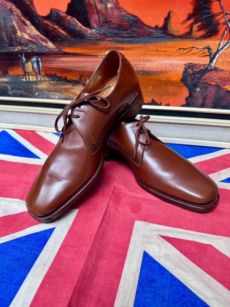 May include: A pair of brown leather dress shoes with laces. The shoes have a square toe and are displayed on a Union Jack flag. The background features a framed landscape painting with a sunset scene.