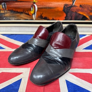 K&ouml;nnte beinhalten: Ein Paar graue Lederschuhe mit burgunderfarbenem Blatt und schwarzen und grauen Akzenten. Die Schuhe werden auf einer Union Jack-Flagge mit einem Gem&auml;lde im Hintergrund pr&auml;sentiert.