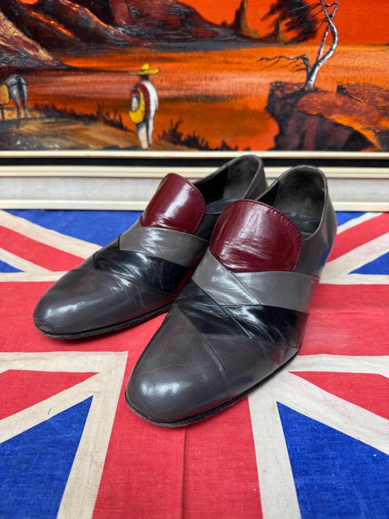 K&ouml;nnte beinhalten: Ein Paar graue Lederschuhe mit burgunderfarbenem Blatt und schwarzen und grauen Akzenten. Die Schuhe werden auf einer Union Jack-Flagge mit einem Gem&auml;lde im Hintergrund pr&auml;sentiert.