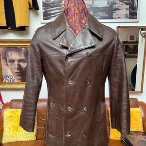 May include: A brown leather double-breasted jacket with a notched collar and four buttons. The jacket has two front pockets and long sleeves. A patterned scarf is draped over the collar. The jacket is displayed on a mannequin.