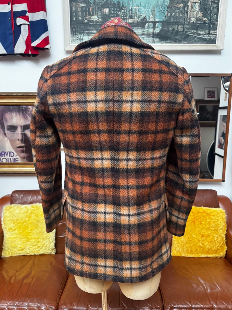 May include: A brown, orange, and black plaid jacket. The jacket is displayed on a mannequin. The jacket has a collared neckline and long sleeves. The jacket is made of a textured fabric.