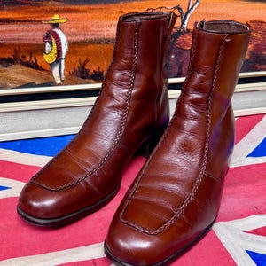 Puede incluir: Botines de cuero marrón con puntera cuadrada y costuras decorativas. Las botas tienen una cremallera lateral. Se muestran sobre una bandera de la Union Jack.