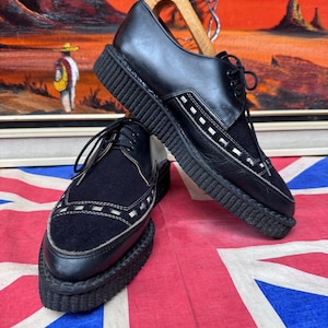 May include: Black leather and suede shoes with thick, ridged soles. The shoes have a classic lace-up design with white stitching details. The shoes are displayed on a Union Jack flag.