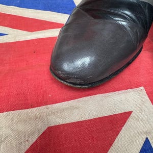 K&ouml;nnte beinhalten: Nahaufnahme eines grauen, schwarzen und dunkelgrauen Lederschuhs. Der Schuh hat eine runde Spitze und ein schwarzes Band &uuml;ber der Oberseite. Der Schuh befindet sich auf einer rot, wei&szlig; und blau gemusterten Oberfl&auml;che.