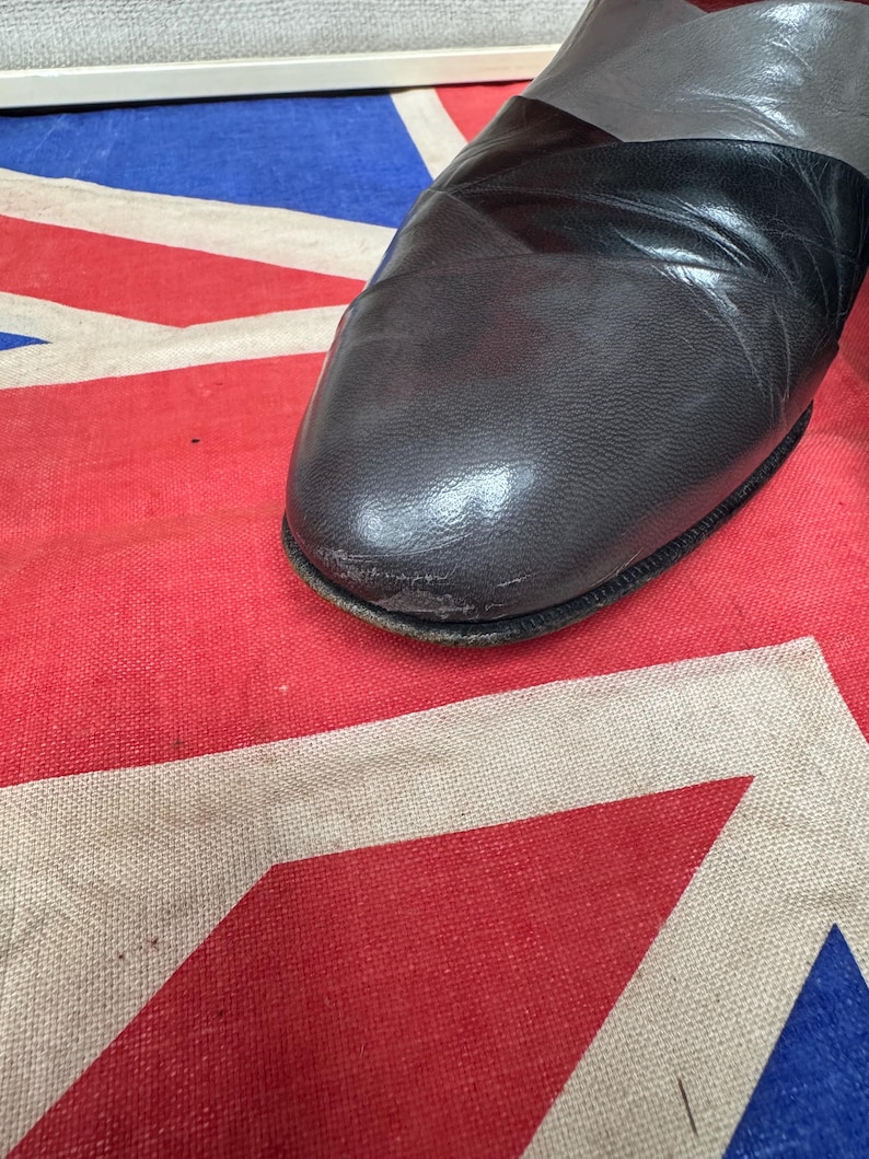 K&ouml;nnte beinhalten: Nahaufnahme eines grauen, schwarzen und dunkelgrauen Lederschuhs. Der Schuh hat eine runde Spitze und ein schwarzes Band &uuml;ber der Oberseite. Der Schuh befindet sich auf einer rot, wei&szlig; und blau gemusterten Oberfl&auml;che.