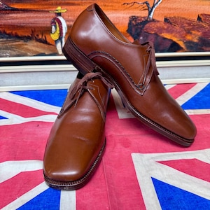 May include: A pair of brown leather dress shoes with laces. The shoes are displayed on a Union Jack flag. The background features a painting of a desert landscape with a figure in the distance.