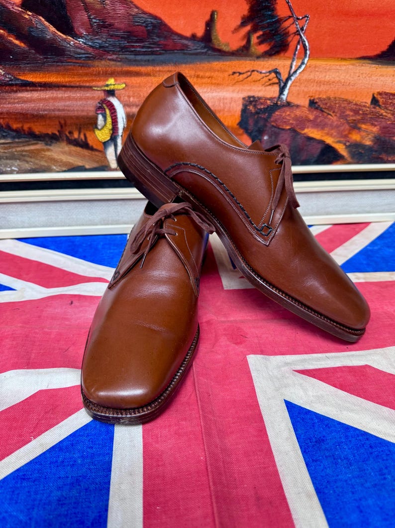 May include: A pair of brown leather dress shoes with laces. The shoes are displayed on a Union Jack flag. The background features a painting of a desert landscape with a figure in the distance.