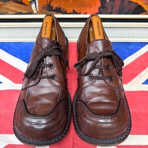 May include: A pair of brown leather shoes with thick soles and dark brown laces. The shoes have a rounded toe design with decorative stitching. Shoe trees are inserted inside the shoes. The shoes are placed on a Union Jack flag.