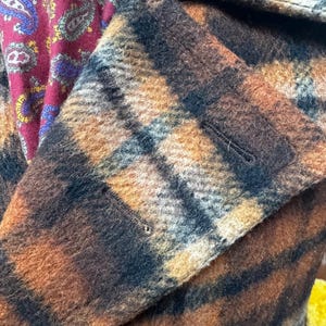 May include: Close-up of a brown and black plaid coat with a buttoned flap. The coat's fabric has a textured appearance. A burgundy paisley patterned fabric is visible in the background.