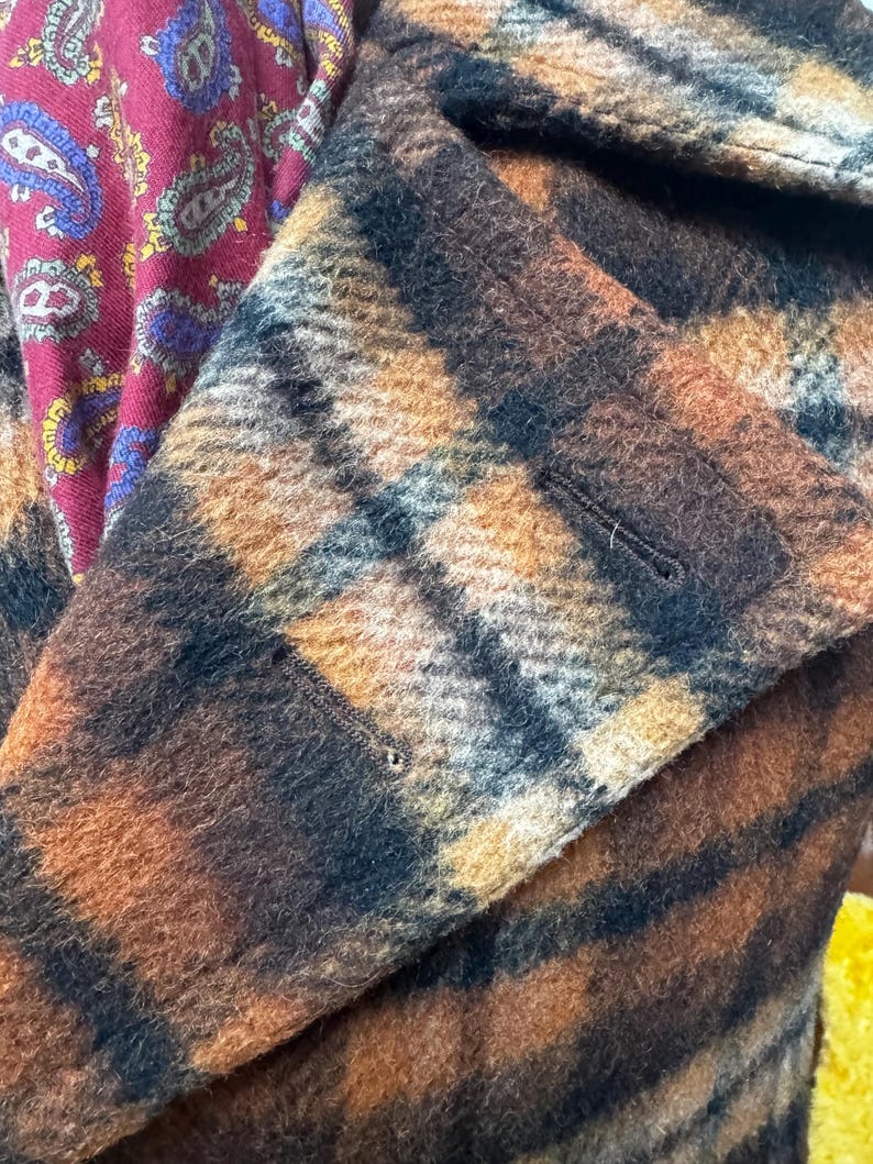 May include: Close-up of a brown and black plaid coat with a buttoned flap. The coat's fabric has a textured appearance. A burgundy paisley patterned fabric is visible in the background.