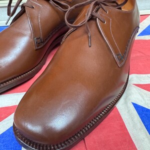 May include: A pair of brown leather dress shoes with laces. The shoes have a classic design with a rounded toe and a stitched sole. The shoes are displayed on a red, white, and blue patterned surface.