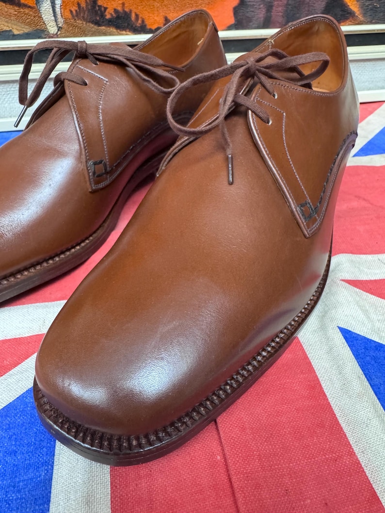 May include: A pair of brown leather dress shoes with laces. The shoes have a classic design with a rounded toe and a stitched sole. The shoes are displayed on a red, white, and blue patterned surface.