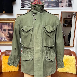 May include: An olive green military-style jacket with multiple pockets and a high collar. The jacket has a button-up front and a drawstring waist. The jacket is displayed on a mannequin. The jacket has a patterned lining visible at the collar.