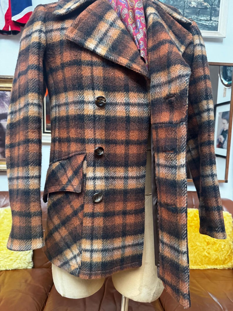 May include: A vintage plaid coat in shades of brown, orange, and black. The coat features a double-breasted design with three buttons, a notched collar, and two front pockets. The coat's interior lining is visible, showcasing a paisley pattern.