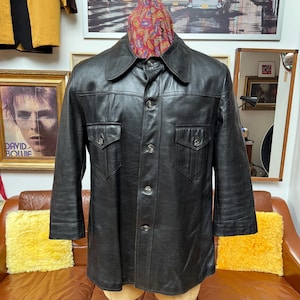 May include: Black leather jacket with a button-up front, two chest pockets, and a collared neckline. The jacket is displayed on a mannequin. A colorful paisley scarf is draped over the mannequin's neck.