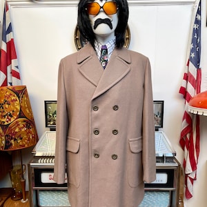 May include: A long, double-breasted, light brown coat with a notched collar and six buttons. The coat has two flap pockets on the front and is worn by a mannequin with a mustache and sunglasses.