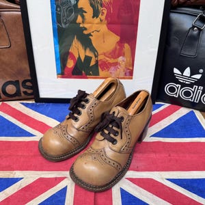 May include: A pair of tan leather heeled shoes with brown laces and decorative perforations. The shoes are displayed on a Union Jack flag, with a framed artwork and leather bags in the background.
