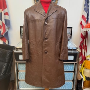 Op de afbeelding: Een bruine leren jas met een enkele rij knopen en een reverskraag. De jas is lang en wordt gepresenteerd op een mannequin.