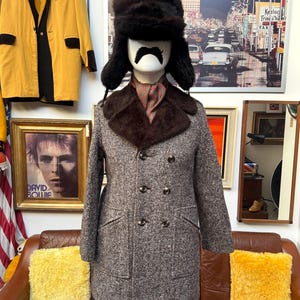 Puede incluir: Un abrigo de tweed vintage con cuello de piel marrón y botones de doble botonadura. El maniquí lleva un gorro de piel y una bufanda estampada. Una blazer dorada con ribete negro cuelga en el fondo, junto a un retrato enmarcado de David Bowie.