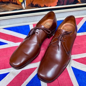 May include: A pair of brown leather dress shoes with laces, displayed on a Union Jack flag. The shoes have a classic design with a rounded toe and stitched detailing. The brand name "Hornes" is visible inside the shoe.