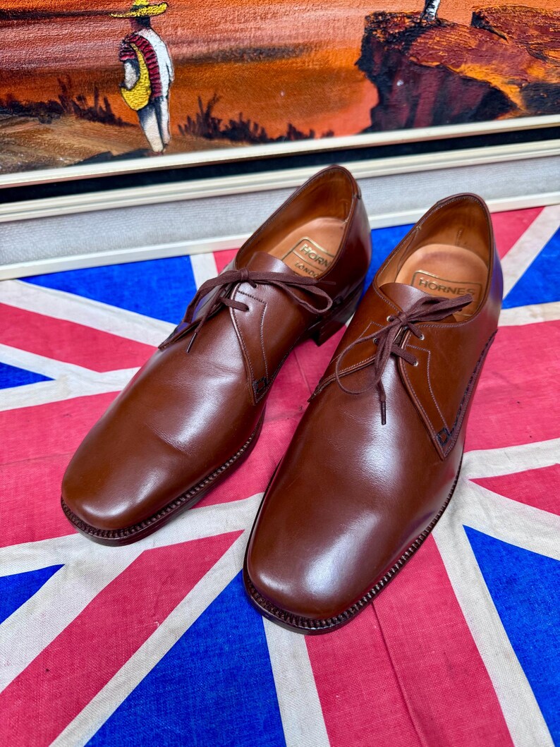 May include: A pair of brown leather dress shoes with laces, displayed on a Union Jack flag. The shoes have a classic design with a rounded toe and stitched detailing. The brand name "Hornes" is visible inside the shoe.