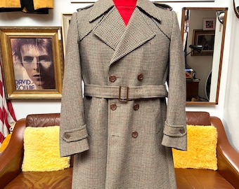 May include: A vintage houndstooth pattern trench coat in brown and tan, double-breasted with a belt. The coat has a classic design with button details and epaulets. The coat is displayed on a wooden stand.